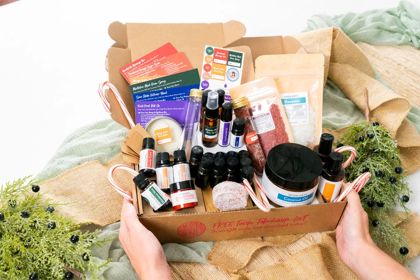 Hands holding the open November Recipe Box, packed with its oils, recipe cards, cranberry seeds and supplies, among juniper and candy canes